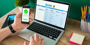 Wire transfer. Young man using his smartphone and laptop to do a banking transaction and some payments